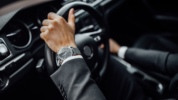 Close up top view of man's watch in black suit keeping hand on the steering wheel while driving a luxury car.
