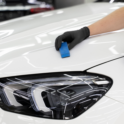 Detailer's gloved hand using a blue clay bar on the hood of a white car to prep the surface for detailing.
