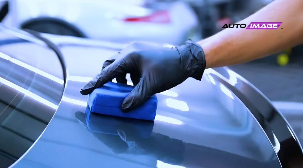 Close-up of a gloved hand applying ceramic coating with a blue applicator pad to a sleek car surface. The finish is highly reflective, capturing light strips and the surrounding environment, with an “Auto Image” logo visible in the upper right.