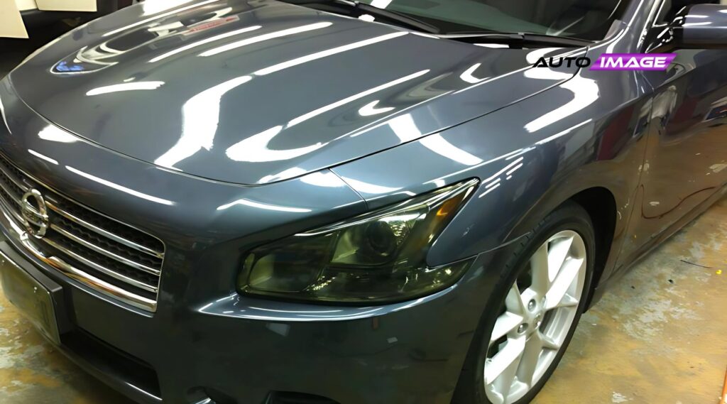 Close-up of a shiny, dark gray Nissan sedan with a reflective hood and headlight, taken indoors at an auto detailing shop. The image features a logo overlay in the top right corner reading "AUTO IMAGE" in bold black and purple text.