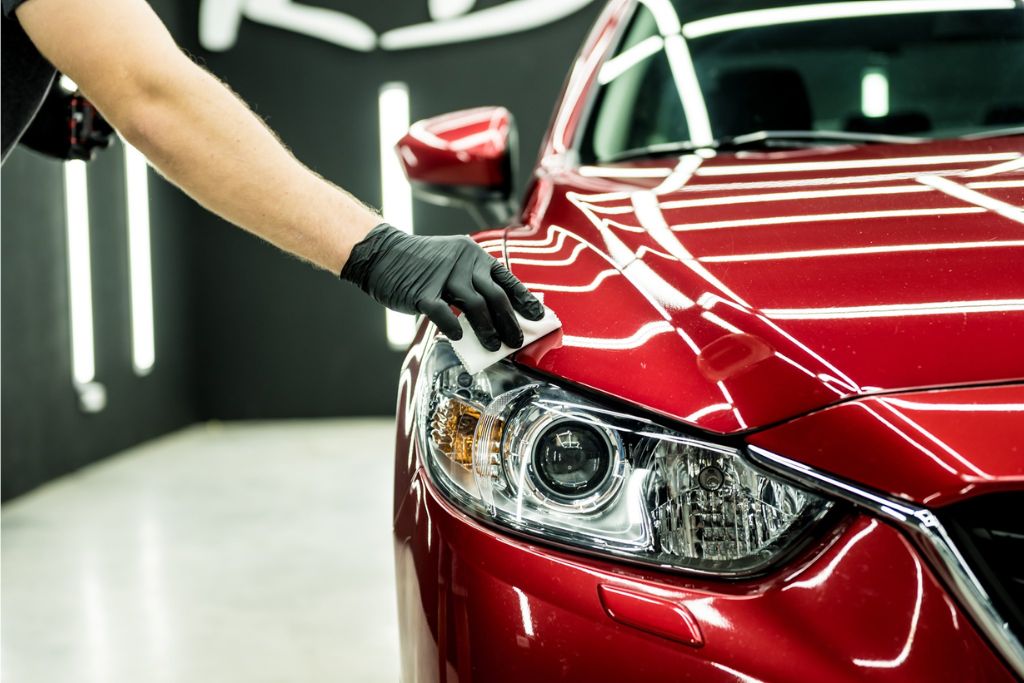 A detailer wearing a black glove applies ceramic coating to the front headlight and fender of a glossy red car under bright, reflective lighting in a professional detailing bay.