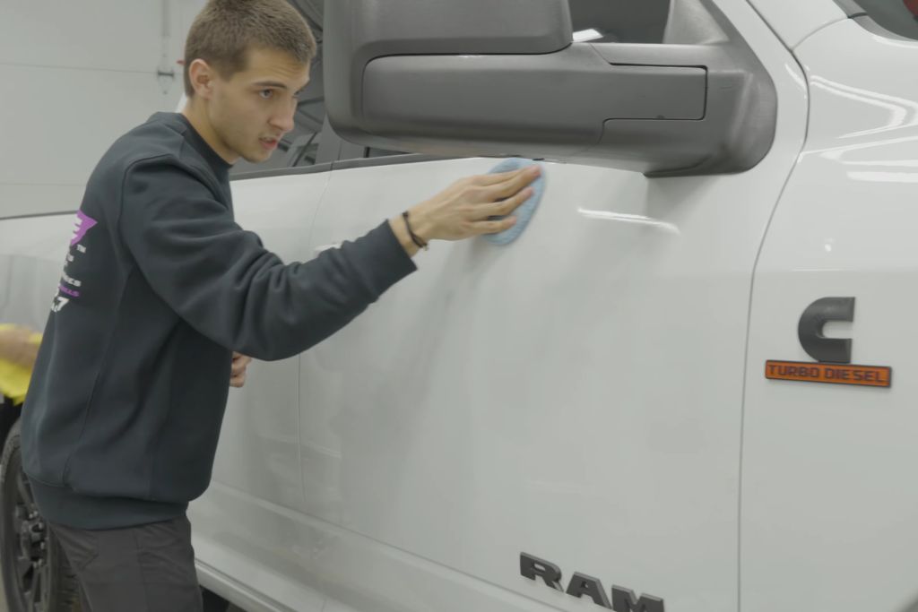 An auto detailing technician buffs the side of a white RAM pickup truck using a soft blue applicator pad, enhancing the finish with ceramic coating. The truck's “Turbo Diesel” badge is visible, emphasizing durability and long-term vehicle care.