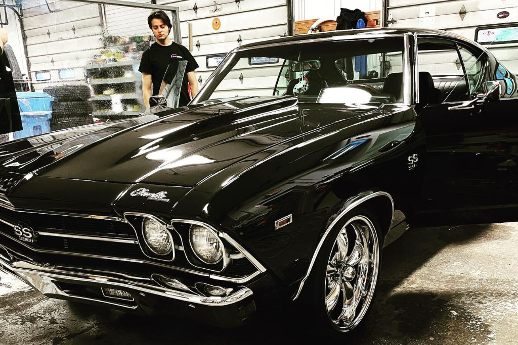 A freshly detailed black Chevy Chevelle SS 396 shines under shop lights, showcasing a deep gloss finish achieved with professional ceramic coating. A technician from Auto Image stands nearby, emphasizing the skill and care involved in expert application.