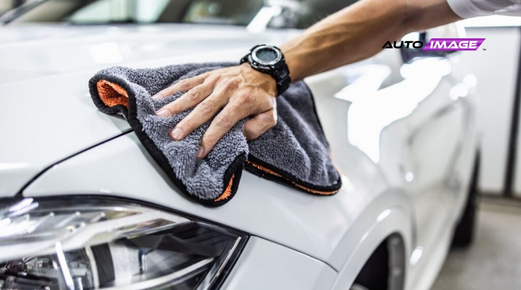 A close-up of a person's hand using a soft microfiber cloth to polish the hood of a clean white car, emphasizing the smooth and glossy surface. The person wears a black digital wristwatch, and the "Auto Image" logo appears in the background, suggesting professional car detailing.