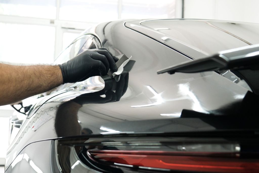 Close-up of a gloved hand applying ceramic coating to the rear surface of a glossy black car, illustrating the precise, professional technique used to create a durable, hydrophobic layer that protects the paint.