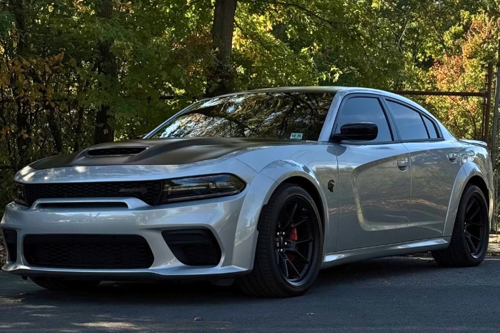 A sleek silver Dodge Charger with a matte black hood is parked on a shaded road, its glossy ceramic-coated exterior reflecting the surrounding trees. The car’s clean surface and deep shine highlight the benefits of ceramic coating for long-lasting gloss and protection.