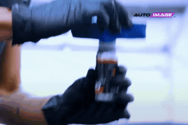 Close-up of a gloved technician applying ceramic coating from a small bottle onto an applicator pad, with the "Auto Image" logo in the corner. The motion highlights the care and precision used during the coating process.