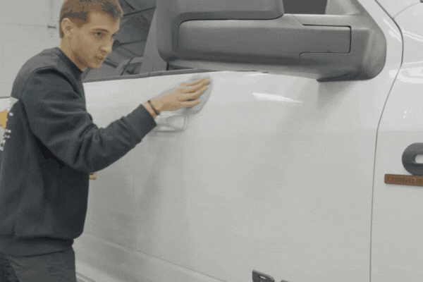 A detailing professional gently buffs the door of a white truck using a microfiber cloth, smoothing out the ceramic coating for an even, glossy finish. The technician moves with precision, highlighting the care and skill involved in applying the protective layer.