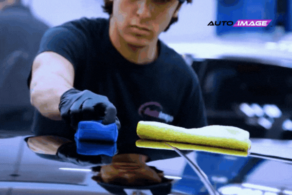 A detailing specialist at Auto Image carefully applies ceramic coating to the roof of a glossy black car using a blue applicator block, with a yellow microfiber cloth resting nearby. The Auto Image logo is visible in the top corner, emphasizing their professional detailing services.
