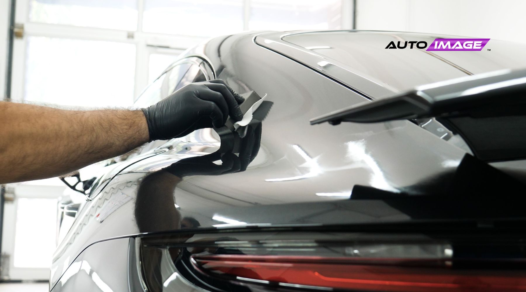 A gloved hand applies a protective ceramic coating to the sleek, glossy surface of a black car, reflecting the lights and surrounding environment; the "Auto Image" logo appears in the top right corner.