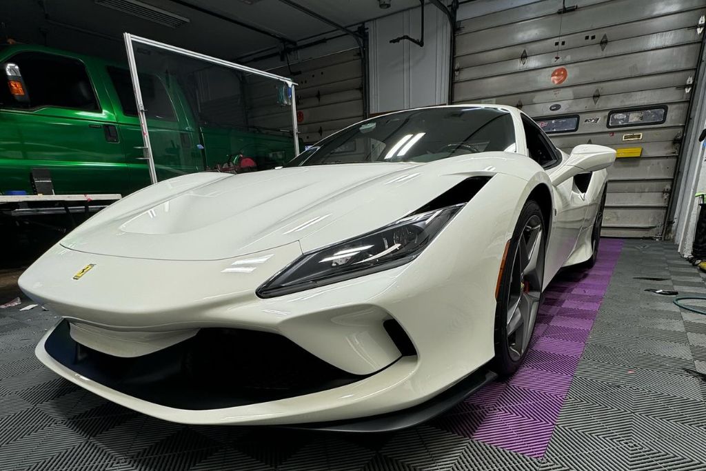 A sleek white Ferrari sports car parked in a professional detailing garage, its glossy finish reflecting overhead lights—highlighting the protective shine likely enhanced by ceramic coating. A green truck is visible in the background, along with tools and garage equipment.