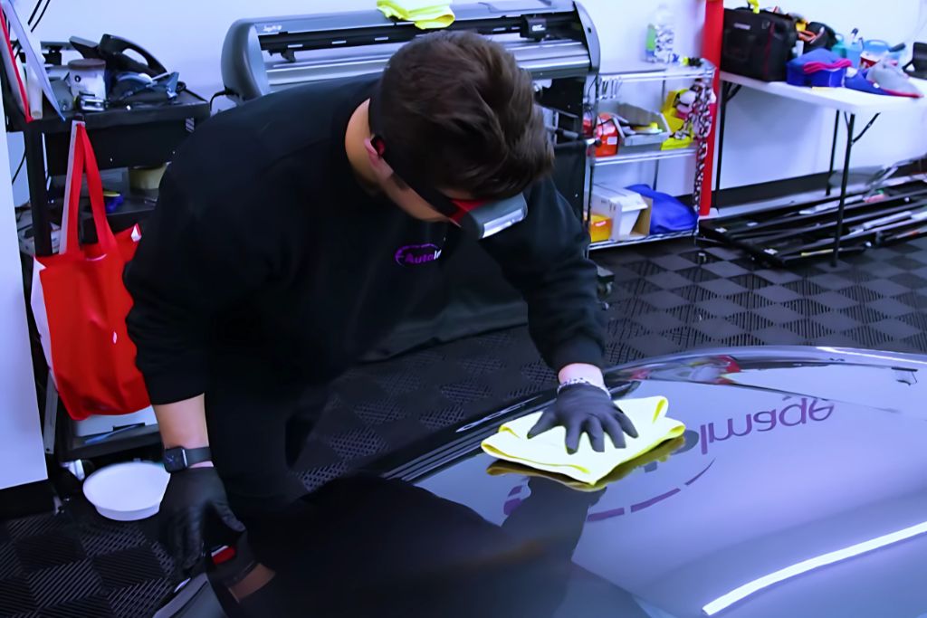 A professional wearing gloves and a headlamp buffs a car hood with a yellow microfiber cloth in a clean, organized auto detailing shop. The reflection on the hood shows the word "Applied," suggesting a focus on ceramic coating or paint protection.