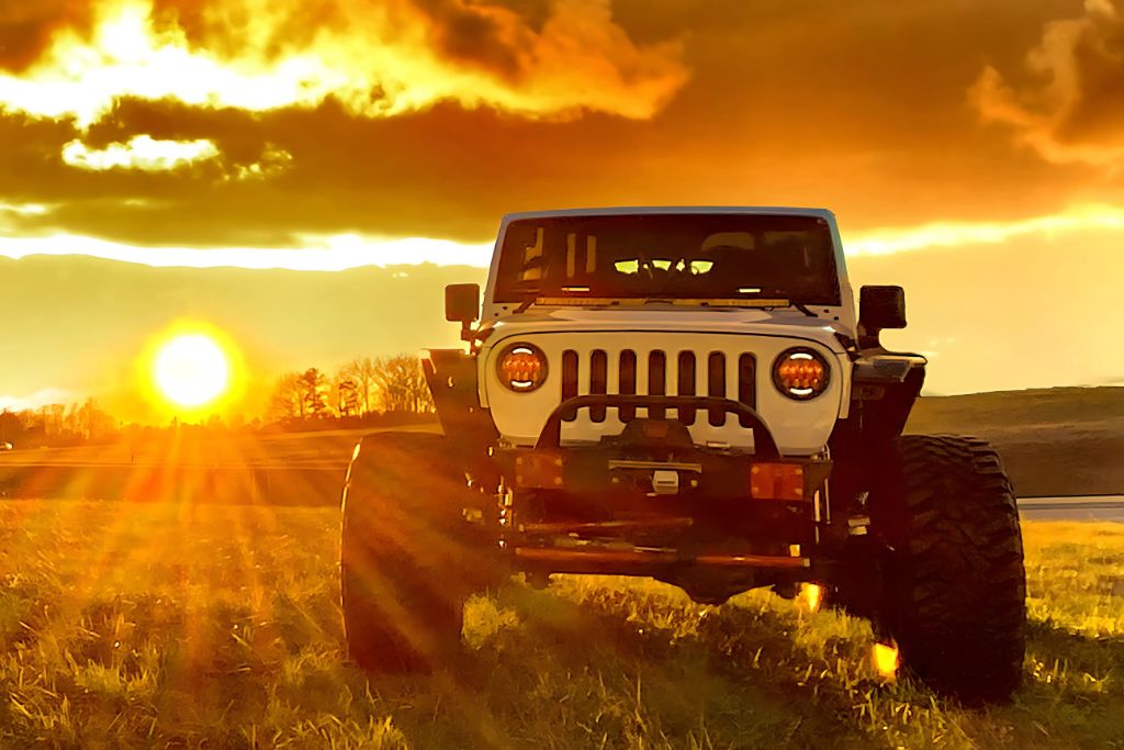 A rugged white Jeep with oversized tires is parked in a grassy field during sunset, with dramatic golden light and sun rays flaring across the image, emphasizing the protective appeal of window tinting against harsh UV rays.