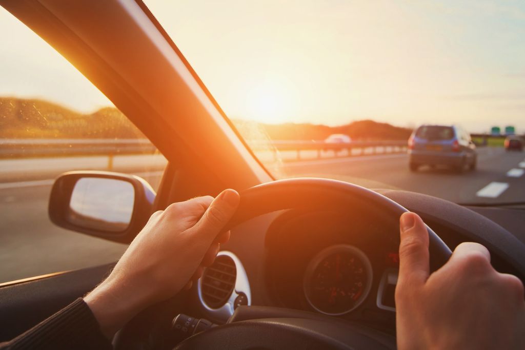 A driver grips the steering wheel while driving into the sunset on a highway, with golden sunlight streaming through the windshield, symbolizing the intense heat window tinting can help reduce.