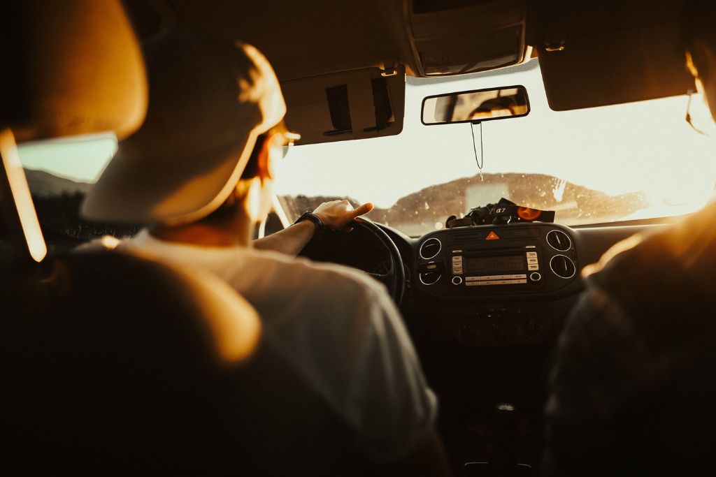 View from the backseat of a car as a driver wearing a cap steers into a glaring sunset, with intense golden light flooding through the windshield and casting shadows across the interior.