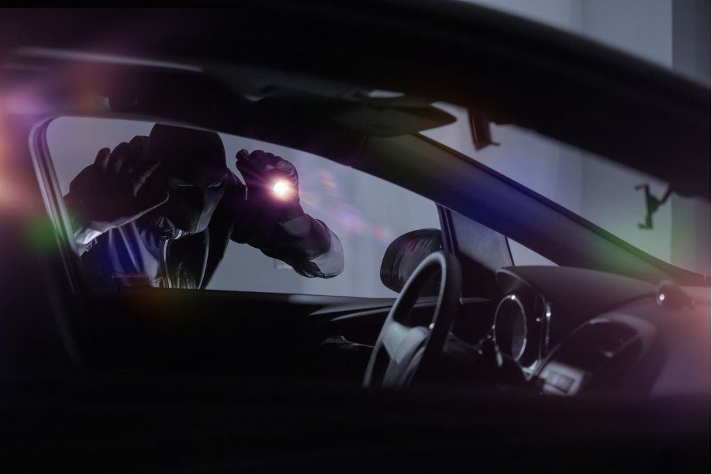 A hooded figure peers with a flashlight into the window of a parked car at night, emphasizing the vulnerability of untinted windows to prying eyes and potential theft.