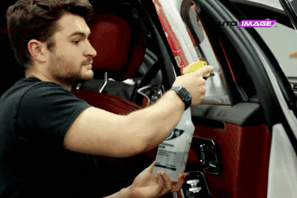 A professional technician sprays cleaning solution onto a car window during a tinting preparation at Auto Image, with a glimpse of the vehicle’s luxurious red interior and the company logo in the corner.