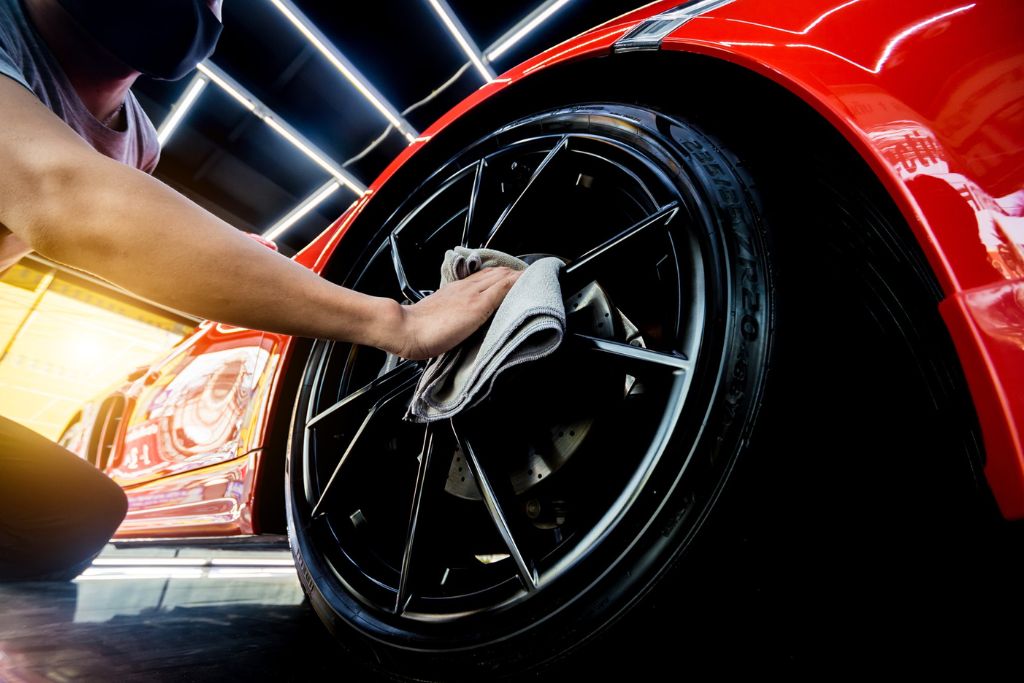 A detailing technician wearing a mask cleans a glossy black wheel on a bright red sports car using a microfiber cloth under sleek LED lighting.