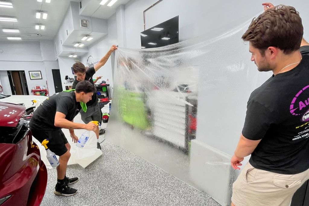 Three technicians in a garage carefully position a large, wet sheet of paint protection film (PPF), preparing it for installation on a vehicle. One holds a spray bottle while others stretch and align the clear film, showcasing the teamwork and precision involved in PPF application.