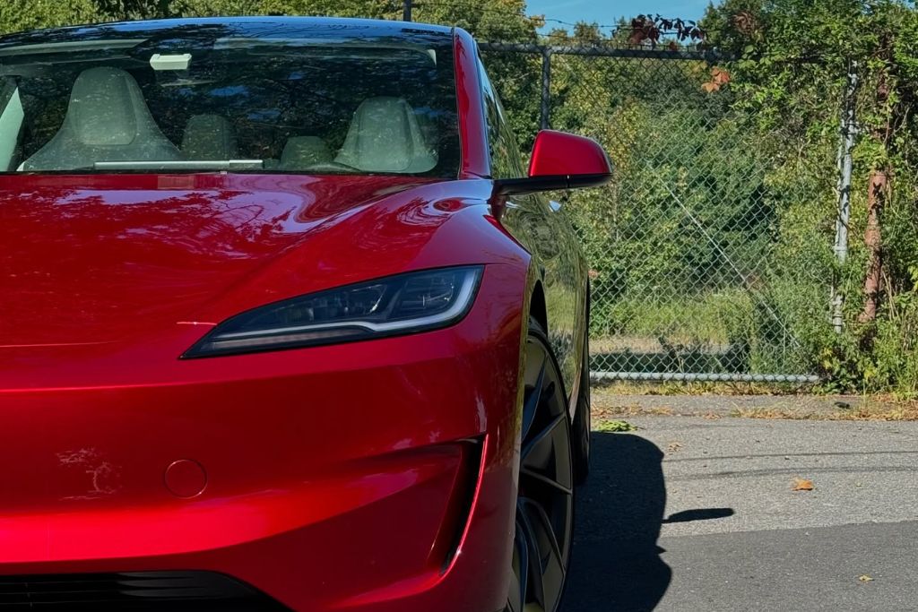 Front view of a glossy red Tesla parked in sunlight, showcasing a flawless paint finish and subtle reflections on the body—suggesting recent paint protection film (PPF) application. The image emphasizes the aesthetic and protective value of PPF for high-end vehicles.