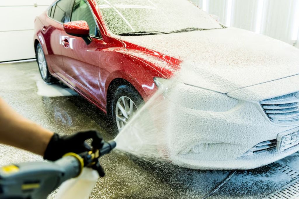 A red sedan is being sprayed with thick white foam during a professional car wash, with half the car already coated. A person in gloves holds the foam cannon, emphasizing the importance of gentle, thorough cleaning in maintaining paint protection film.