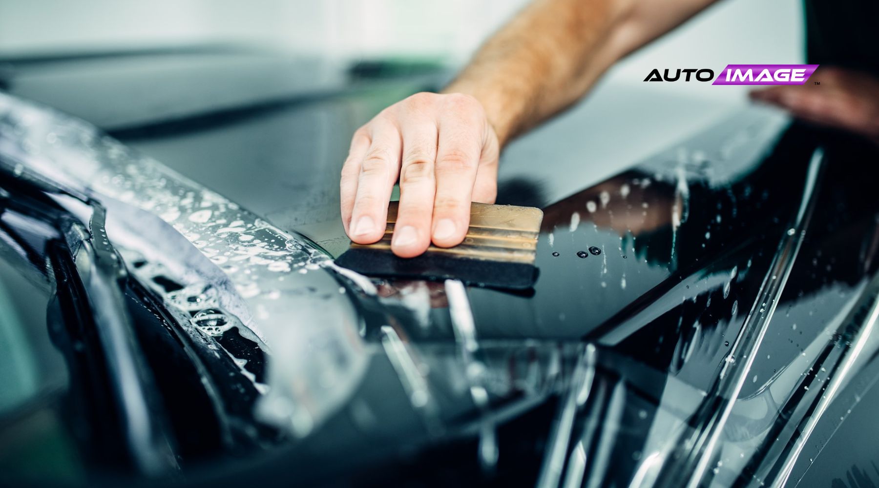 Close-up of a person's hand applying paint protection film (PPF) to a shiny black car hood using a squeegee, with soap suds visible on the surface. The image captures the detailing process, emphasizing the clear, glossy finish and protective care.