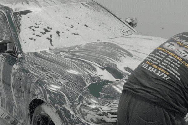 A detailer wearing a branded shirt thoroughly hand-washes a foam-covered car during an in-depth professional car detailing service.