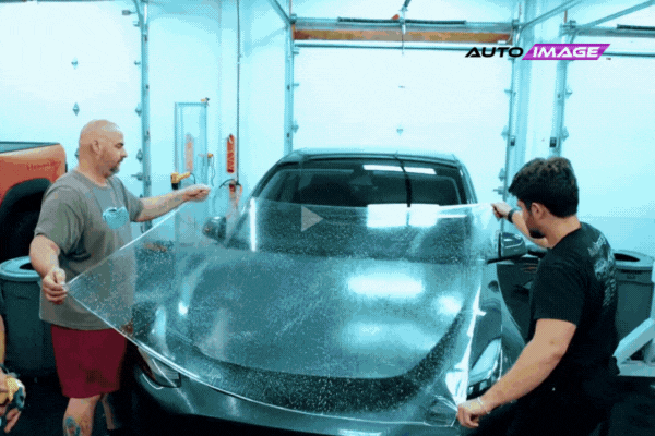 Two Auto Image technicians apply a large sheet of transparent paint protection film (PPF) to the hood of a sleek black sports car inside a clean, well-lit garage. The motion captures their teamwork and precision, emphasizing professional installation quality.