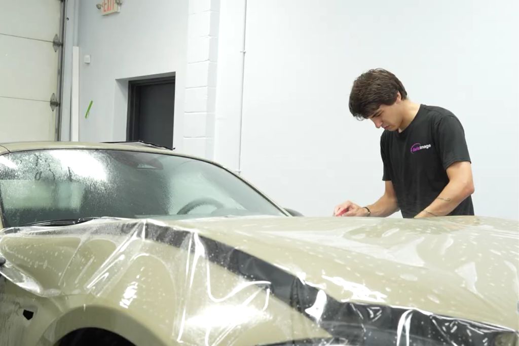 A technician from Auto Image applies paint protection film to the hood of a beige car inside a detailing bay. The transparent film is visibly stretched over the vehicle’s wet surface, showcasing the precision and care involved in a professional PPF installation.