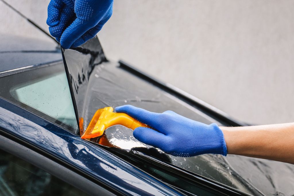 Installer wearing blue gloves applies a sheet of dark 3M window film to a car window using a yellow squeegee tool, ensuring smooth, bubble-free adhesion for long-lasting performance and heat rejection.