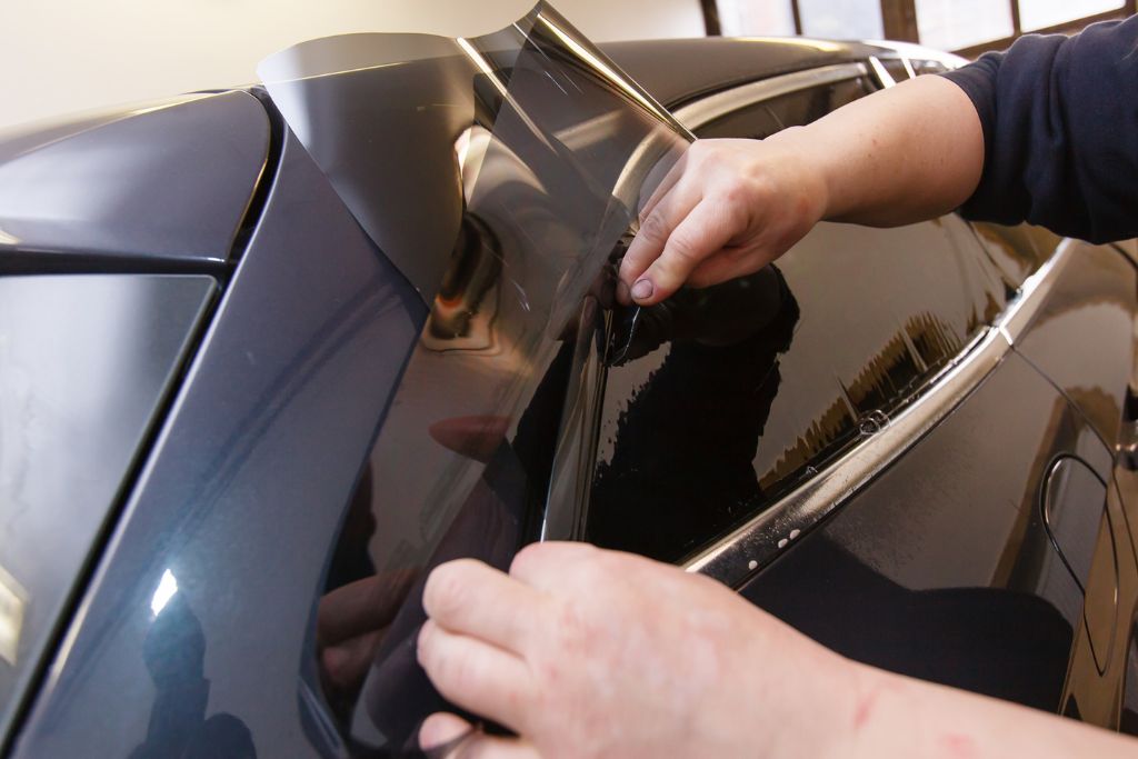 Close-up of hands applying a sheet of 3M Crystalline window film to the rear side window of a Tesla, showing the precise installation process that enhances heat rejection, UV protection, and clarity without affecting signal reception.
