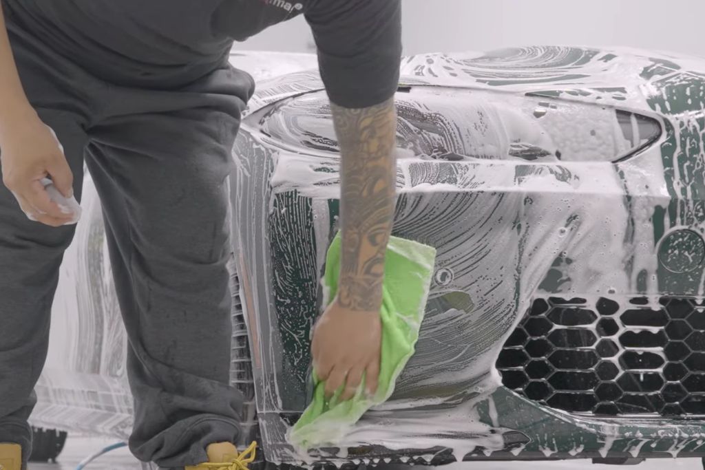Auto detailing technician washing a car’s front bumper with thick foam and a green microfiber cloth as part of the proper prep before coating, ensuring the surface is decontaminated before ceramic application.