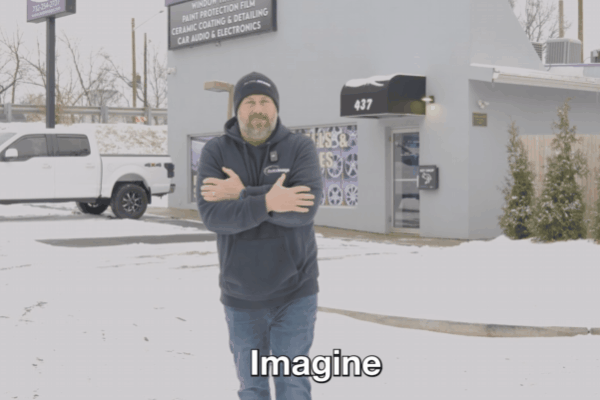 Man bundled in winter clothes shivering with arms crossed, standing outside on a snowy day in front of a building labeled "Paint Protection Film, Ceramic Coating & Detailing, Car Audio & Electronics," with the word "Imagine" captioned below him.