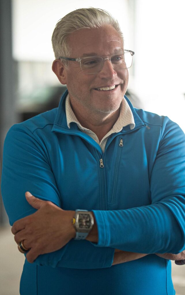 Ryan Warner, a middle-aged man with short silver hair and glasses, smiles confidently with arms crossed while wearing a bright blue zip-up jacket over a patterned white shirt. His polished look, including a silver watch and wedding band, reflects his professional yet approachable leadership style.