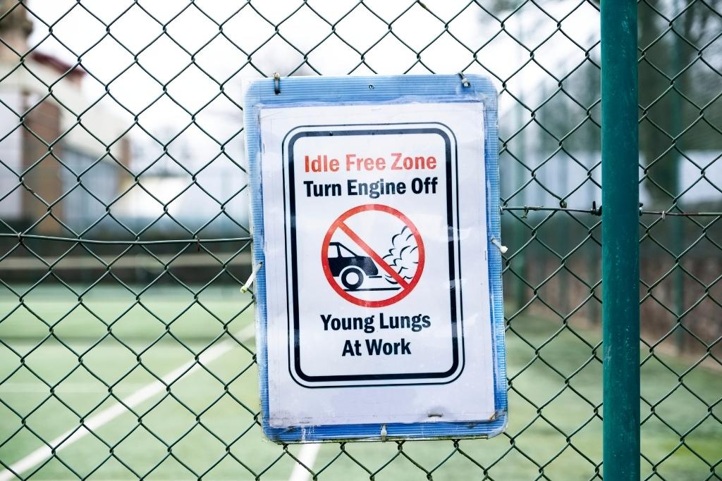 A sign attached to a chain-link fence that reads “Idle Free Zone – Turn Engine Off – Young Lungs At Work,” featuring a crossed-out idling car icon.
