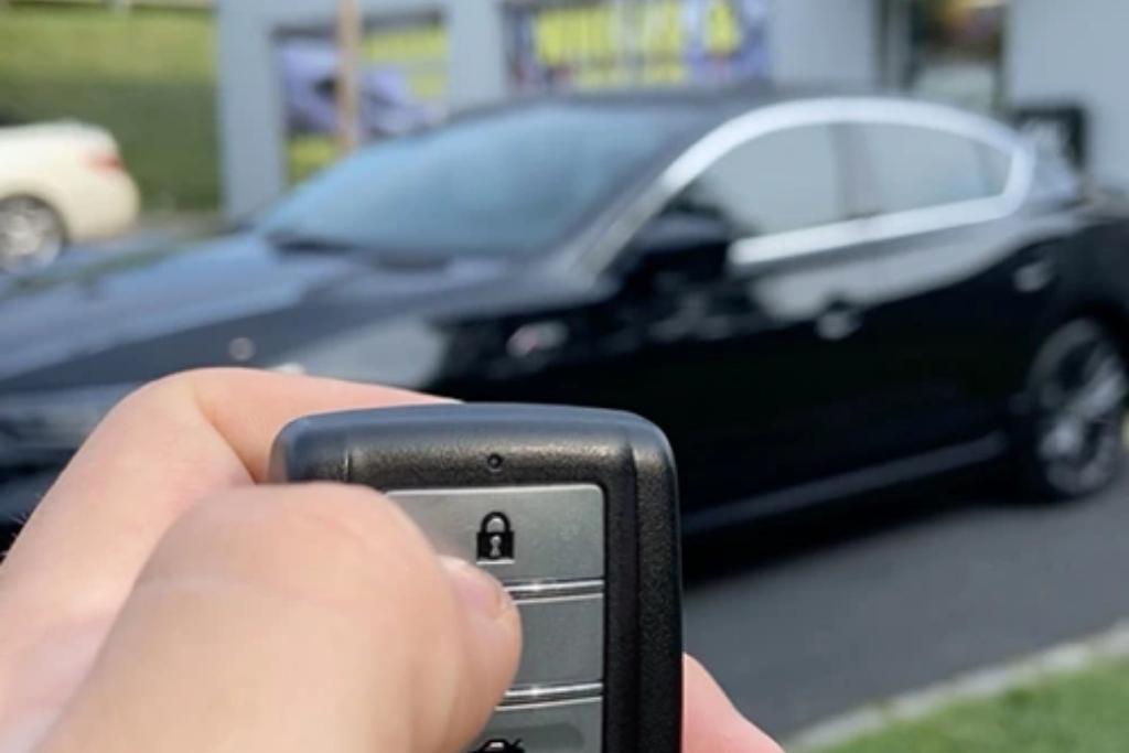 A close-up of a person pressing the lock button on a car remote key while a black sedan with tinted windows is parked in the background.