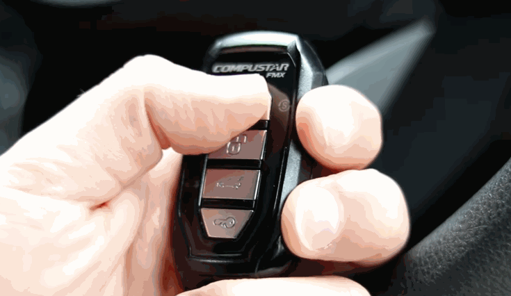 Thumb pressing a button on a Compustar remote start key fob with labeled lock, trunk, and start buttons.