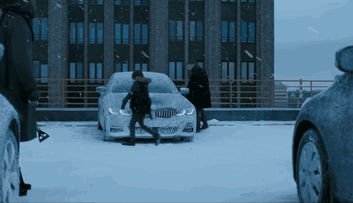 Two people approaching a snow-covered BMW in a snowy parking lot with snow falling.