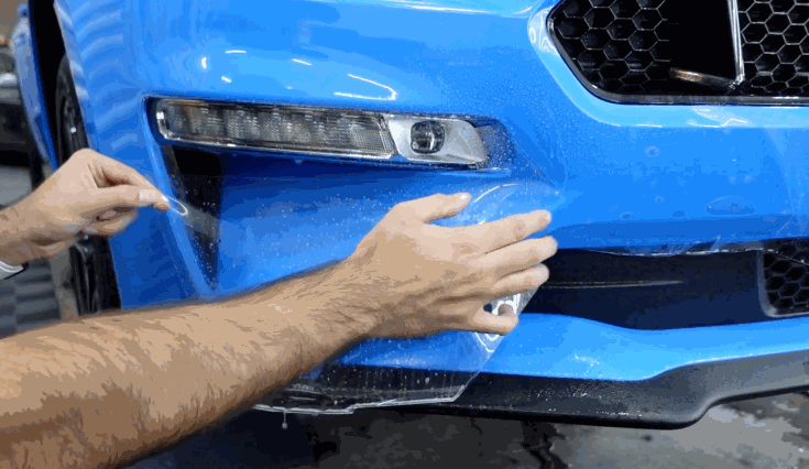 Person applying transparent paint protection film to the bumper of a blue car, with water droplets on the surface.
