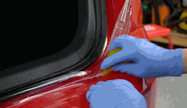 Gloved hands applying clear paint protection film to the edge of a red vehicle near the trunk using a yellow squeegee.