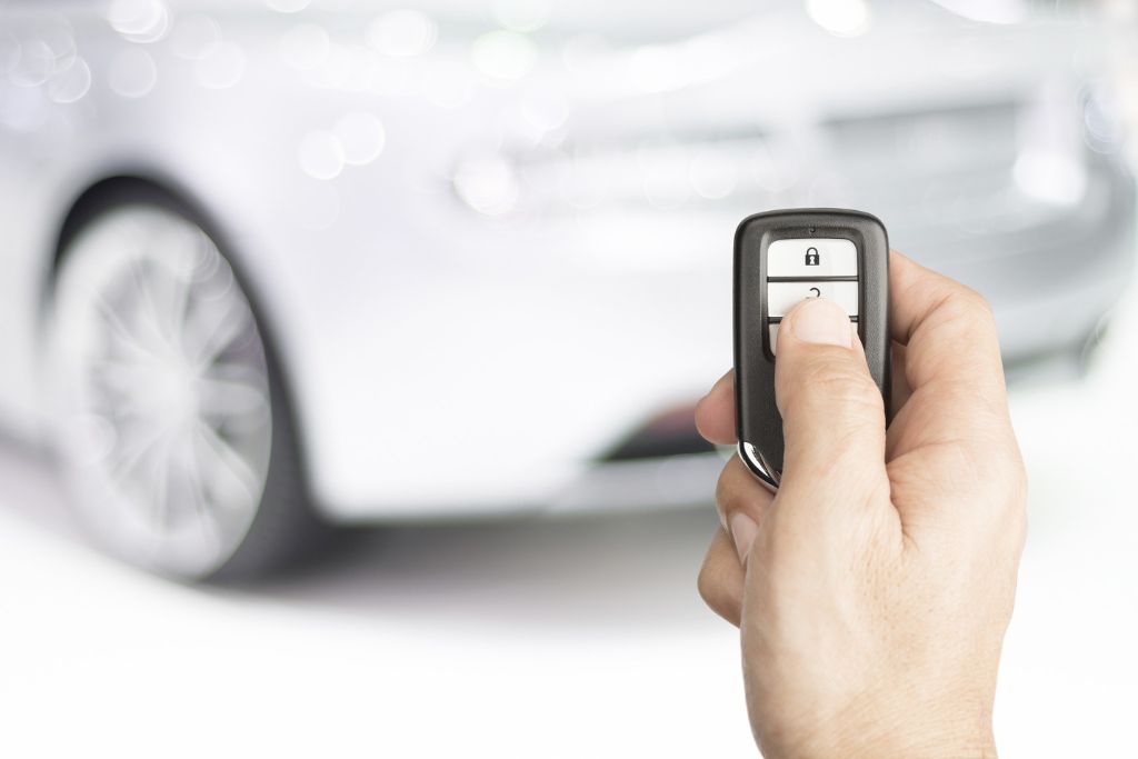 Close-up of a hand holding a car remote starter key fob with a blurred silver car in the background. The thumb is pressing the lock button, symbolizing remote start functionality for vehicles.