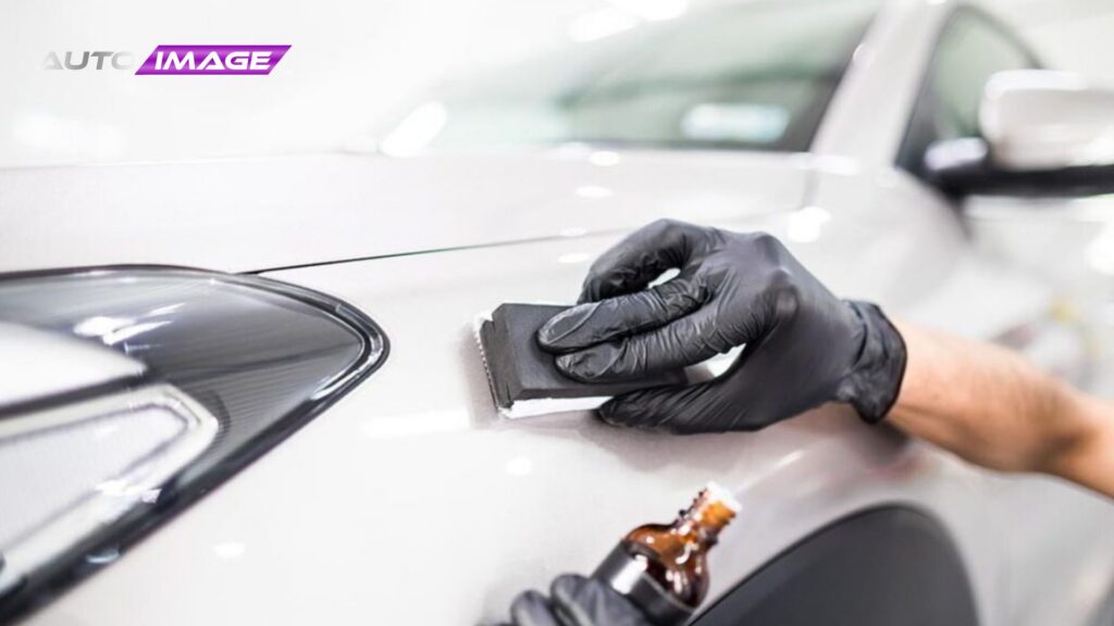 Gloved technician applies ceramic coating to a white vehicle using a small applicator block while holding a bottle of coating solution nearby. The product is carefully spread in controlled strokes to ensure even coverage and durable paint protection.