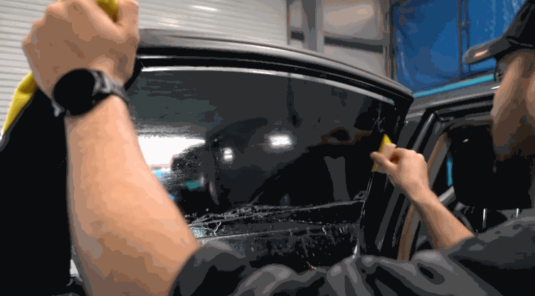 Technician carefully applies Window Film to the side window of a Tesla inside a garage. He smooths the dark tinted film across the glass to remove bubbles and ensure a seamless fit.
