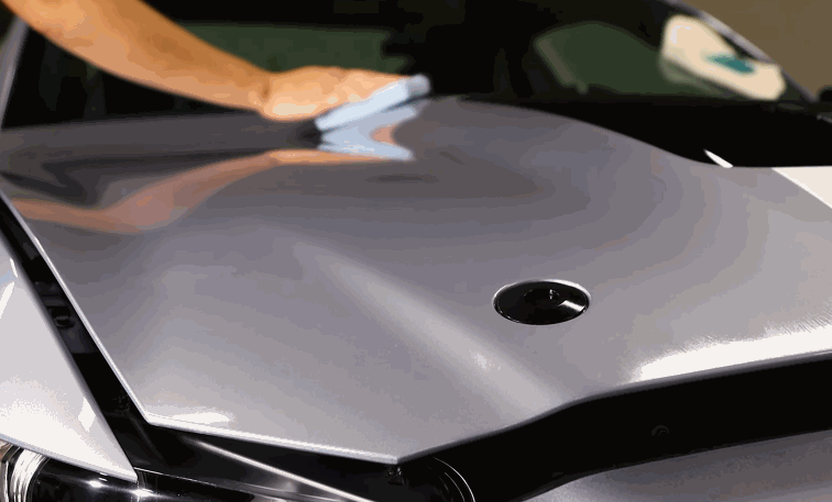 Detailer wipes down a freshly treated hood after ceramic coating application to level and buff the surface. The paint appears smooth and highly reflective under indoor lighting.