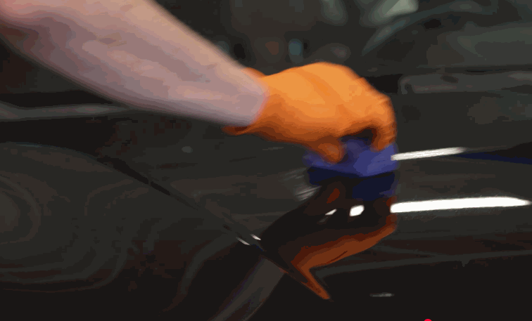 Technician wearing an orange glove applies ceramic coating to a glossy black vehicle panel using a blue applicator pad.