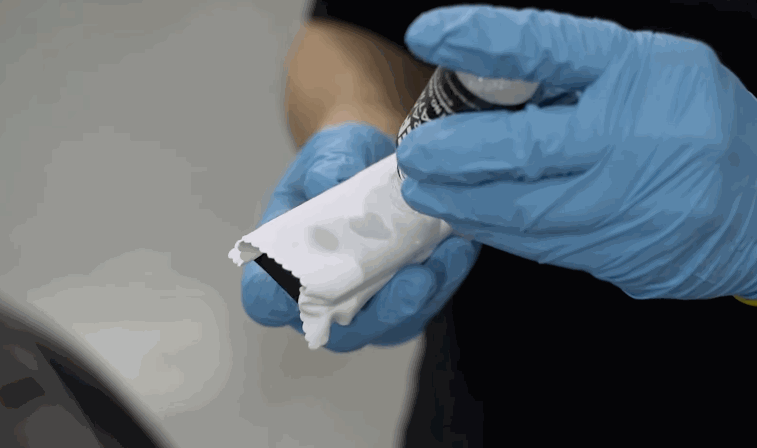 Blue gloved hands hold a folded microfiber cloth while dispensing liquid ceramic coating from a small bottle onto the fabric. The applicator cloth is prepared before spreading the coating across the vehicle paint surface.