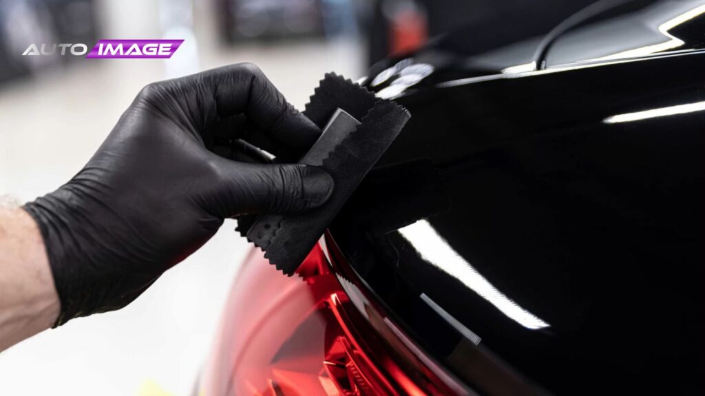Technician wearing a black glove wipes the edge of a glossy black car panel near a red tail light using a small microfiber cloth while applying ceramic coating.