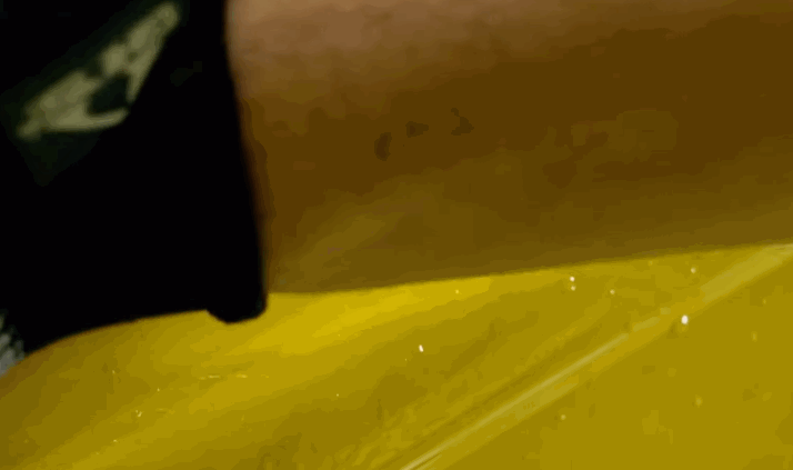 A sheet of transparent paint protection film being laid across a bright yellow car panel while a technician smooths the surface. Small water droplets sit between the film and the paint as the installer positions the protective layer.