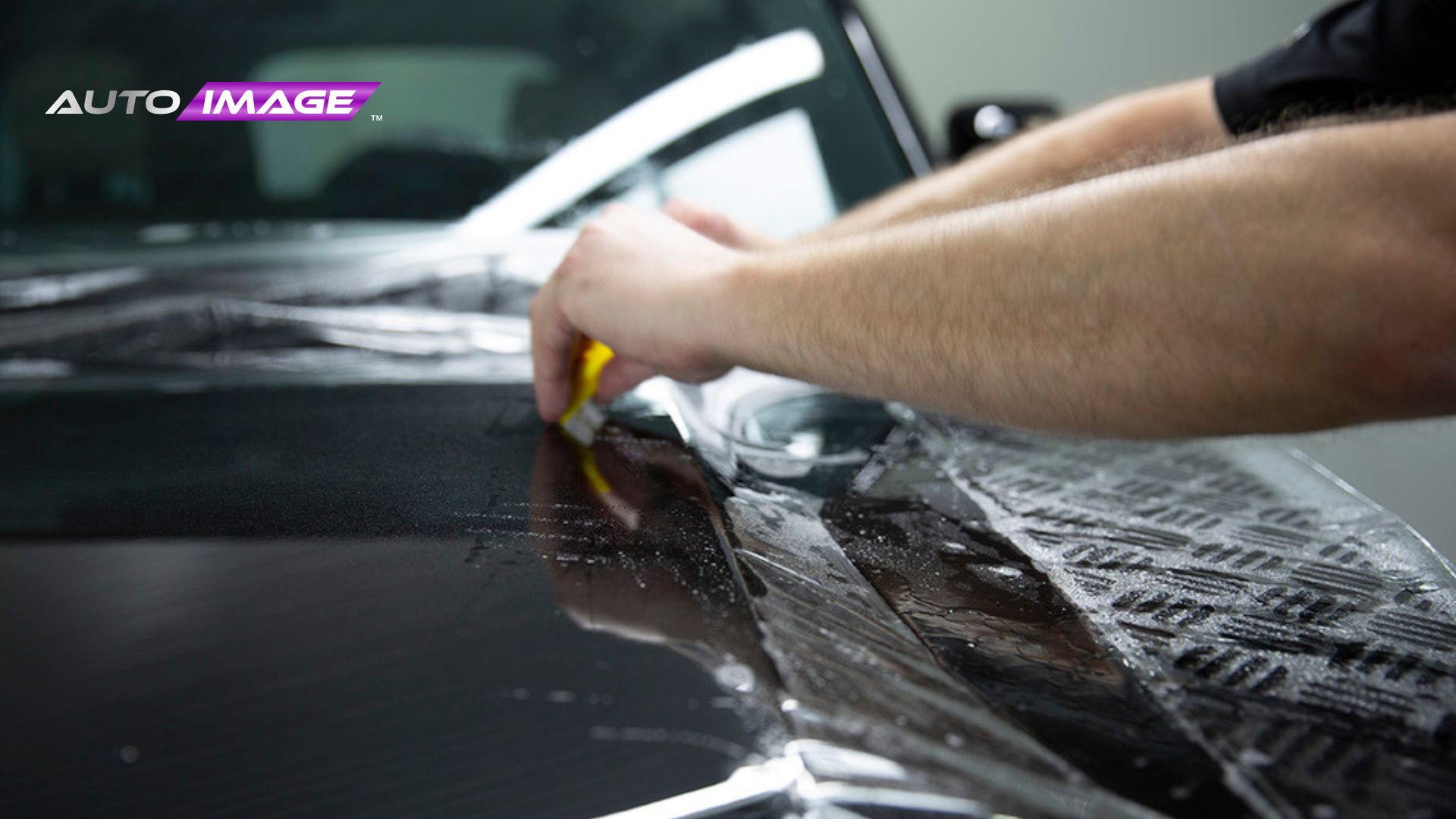 A technician uses a yellow squeegee to press water out from under paint protection film on a black car hood while installing the protective layer. The logo "AUTO IMAGE" appears in the upper left corner as the film is carefully smoothed across the paint.