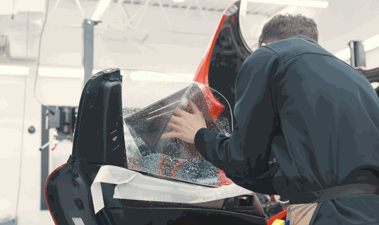 Installer uses both hands to smooth window tint film onto a car window covered in water solution inside a workshop. The scene highlights the step by step process of window tint application for a seamless result.